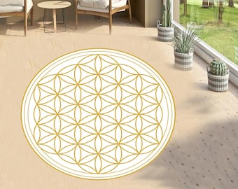 Op de afbeelding: Een ronde, witte vloerkleed met een goudkleurig Flower of Life-patroon. Het kleed ligt op een lichte houten vloer, met stoelen en een kleine tafel op de achtergrond. De kamer heeft een groot raam met uitzicht op bomen.
