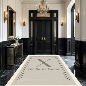 以下が含まれることがあります： 黒い縁取りとグレーの「X」と「The Xavier Family」の文字が入った長方形のベージュ色のウェルカムマット。 黒いドア、白い壁、シャンデリアのある壮大な玄関にマットが置かれています。