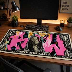 May include: A rectangular desk mouse mat featuring a stylized design of a one-dollar bill with graffiti-style pink lettering. The design includes a portrait of George Washington and the words "ONE DOLLAR".