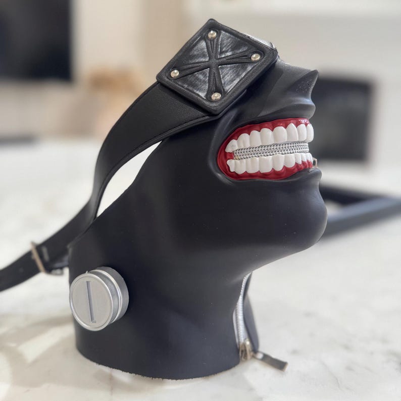 K&ouml;nnte beinhalten: Schwarze Maske mit rotem Mund und wei&szlig;en Z&auml;hnen. Die Maske hat ein schwarzes Band und ein silbernes Augenpflaster mit einem Kreuzmuster.