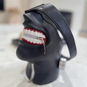 K&ouml;nnte beinhalten: Schwarze Tasche mit Rei&szlig;verschluss in Form eines menschlichen Kopfes mit offenem Mund, der wei&szlig;e Z&auml;hne und rotes Zahnfleisch zeigt. Die Tasche hat einen schwarzen Lederriemen.