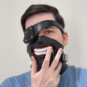 K&ouml;nnte beinhalten: Eine schwarze und rote Maske mit einem Rei&szlig;verschluss-Mund und einem schwarzen Leder-Augenklappen. Die Maske wird von einer Person mit dunklen Haaren und einem blauen Hemd getragen.