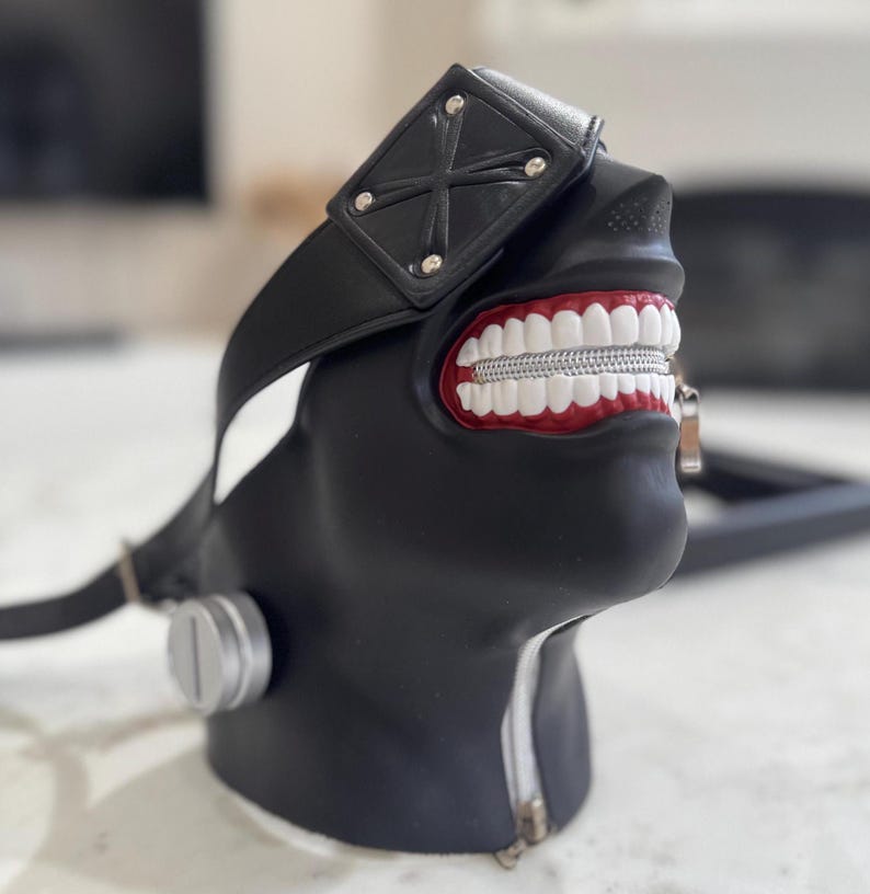 K&ouml;nnte beinhalten: Eine schwarze und rote Maske mit einem Rei&szlig;verschluss-Mund und einem schwarzen Band. Die Maske hat ein wei&szlig;es Kreuz auf der Stirn.