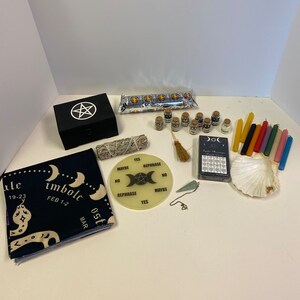 May include: A collection of spiritual and metaphysical items, including a black box with a pentagram, a Ouija board, candles, incense, small jars, a seashell, and a cloth with moon phases and the word "imbolc".