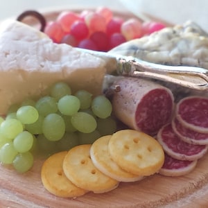 Puede incluir: Una tabla de queso de madera redonda con una variedad de quesos, uvas, galletas y salami. La tabla es de madera natural.