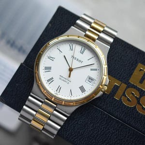 May include: A Tissot Seastar Quartz wristwatch with a white dial, black Roman numeral hour markers, and a date window at the three o'clock position. The watch features a two-tone silver and gold-colored metal link bracelet and a gold-tone bezel.