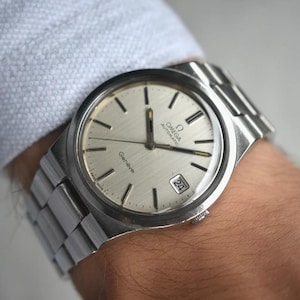 May include: A silver-toned Omega Genève automatic wristwatch featuring a round brushed silver dial with black baton hour markers, silver hands, a date window at the three o'clock position, and a stainless steel link bracelet with a folding clasp.