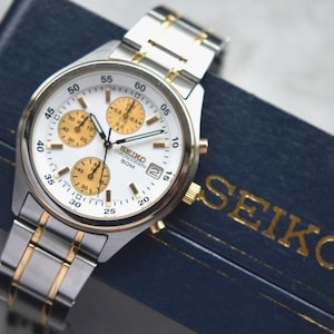 May include: A silver and gold Seiko chronograph watch with a white face and three sub-dials. The watch has a stainless steel strap and is displayed on a blue box with the brand name "SEIKO" in gold lettering.