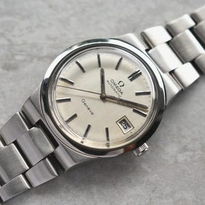 Pode incluir: Um relógio de pulso automático Omega Genève prateado com mostrador redondo em tom off-white, índices de bastão prateados e janela de data na posição das três horas. O relógio possui uma pulseira de elos de aço inoxidável com acabamento escovado.