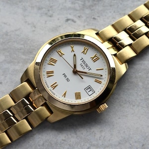 May include: A gold-tone Tissot 1853 PR 50 wristwatch with a white dial, gold Roman numeral hour markers, gold hands, and a date window at the three o'clock position. The watch features a matching gold-tone metal link bracelet and is marked Swiss Made.