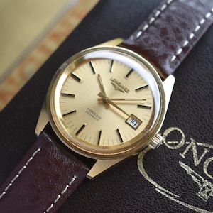 May include: A gold-tone Longines Conquest Automatic wristwatch with a champagne-colored dial, gold baton indices, and a date window at the three o'clock position. The watch features a dark brown textured leather strap with white contrast stitching.