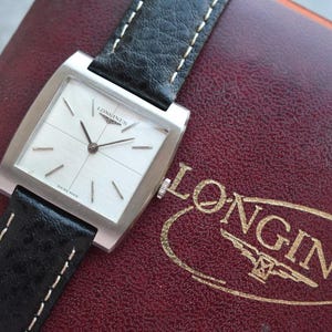 Könnte beinhalten: Eine Vintage-Longines-Uhr mit quadratischem, silberfarbenem Zifferblatt und schwarzem Lederarmband. Die Uhr hat silberne Zeiger und Markierungen, und die Worte "SWISS MADE" sind sichtbar. Die Uhr liegt auf einem weinroten Hintergrund.
