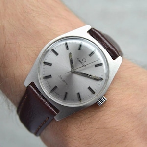 May include: A vintage Omega Genève wristwatch featuring a round silver dial with black baton hour markers, silver hands, and the Omega logo. The watch has a stainless steel case and a dark brown leather strap with visible stitching.