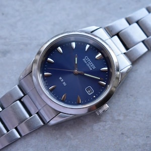 Peut inclure: Une montre Citizen Eco-Drive argentée avec un cadran bleu et des aiguilles argentées. La montre a un affichage de la date à 3 heures et un bracelet argenté. Le cadran de la montre comporte des index argentés. La montre est étiquetée "WR 50".