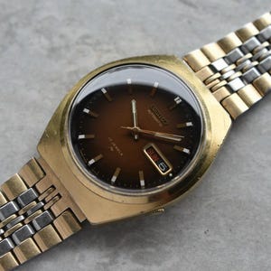Op de afbeelding: Een Seiko automatisch polshorloge met een ronde bruine gradiënt wijzerplaat, goudkleurige wijzers en uurmarkeringen. Het horloge heeft een dag-datumvenster op de drie-uurpositie en een tweekleurige gouden en zilveren metalen schakelarmband.