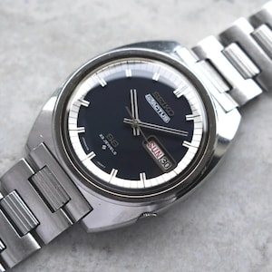 May include: A silver Seiko 5 Actus wristwatch with a dark blue face and a silver strap. The watch face has white hour markers, hands, and a day/date display. The watch is marked "23 Jewels" and "Sun 30".