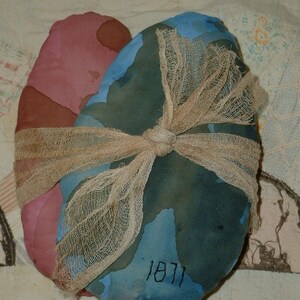 Puede incluir: Dos corazones de tela atados con una cinta de arpillera. Los corazones están pintados en tonos de azul, verde y rosa. El corazón más grande tiene el año 1871 escrito en él.