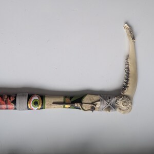 May include: A handcrafted, decorative scythe featuring a bone blade and a wooden handle. The handle is painted with red, orange, and yellow floral patterns, an eye, and geometric designs. The scythe is wrapped with gray twine.