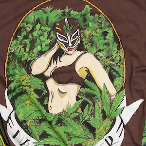 Op de afbeelding: Een bruin T-shirt met een afbeelding van een vrouw die een worstelmasker en een bruine bh draagt, omringd door groene marihuanabladeren. De tekst "Nacho Reefer" staat in wit op een wit lint.
