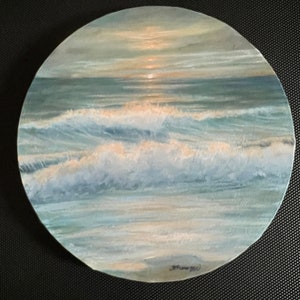 Puede incluir: Pintura redonda de una escena oceánica con olas y una puesta de sol. La obra de arte utiliza tonos de azul, blanco y dorado, capturando la luz sobre el agua. La pintura está firmada en la esquina inferior derecha.
