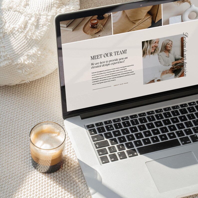 May include: A silver laptop displaying a website with the text MEET OUR TEAM! and Design is our passion. The screen shows images of interior decor and two people working. A glass of coffee with foam sits on a white textured surface next to the laptop.