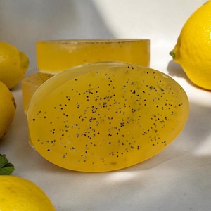 May include: Three oval-shaped bars of yellow soap with black poppy seeds embedded in the soap. The soap is on a white surface with a lemon beside it.