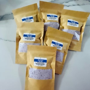 May include: Several kraft paper pouches filled with lavender vanilla bath powder. Each pouch has a blue and white label that reads "Lavender Vanilla Bath Soak Powder". The powder is a light purple color.