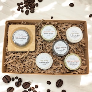 May include: A gift box filled with six round, natural body scrub bars. The scrub bars are in a variety of colors and have labels with different coffee-themed scents: Salted Caramel Liquor, Cinnamon Cappuccino, Vanilla Hazelnut. The box is filled with brown shredded paper and the scrub bars are arranged in a circle.