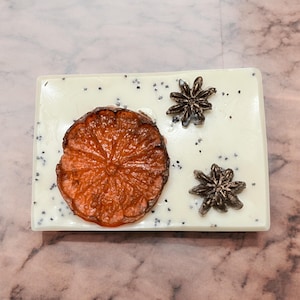 May include: A white wax melt with a dried orange slice, two star anise, and black poppy seeds on top.
