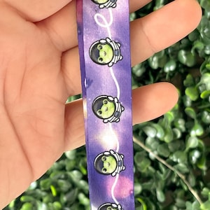 Pu&ograve; includere: Tessuto tie-dye viola e bianco con un motivo ripetuto di rane verdi che indossano caschi da astronauta. Il tessuto ha un anello metallico argentato attaccato alla parte inferiore.