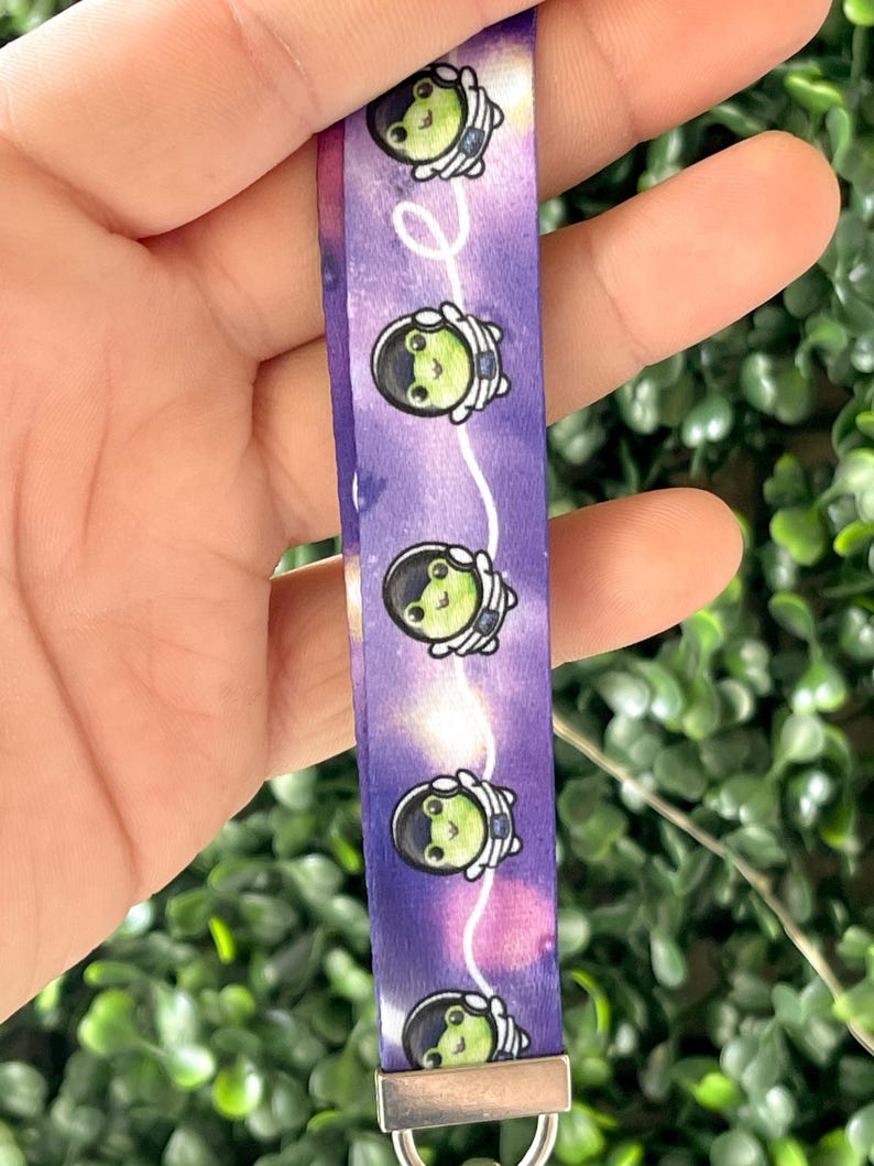 Pu&ograve; includere: Tessuto tie-dye viola e bianco con un motivo ripetuto di rane verdi che indossano caschi da astronauta. Il tessuto ha un anello metallico argentato attaccato alla parte inferiore.