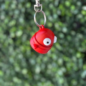 May include: A red, cartoon-like keychain with a single white eye and a small mouth. The keychain is attached to a silver key ring.