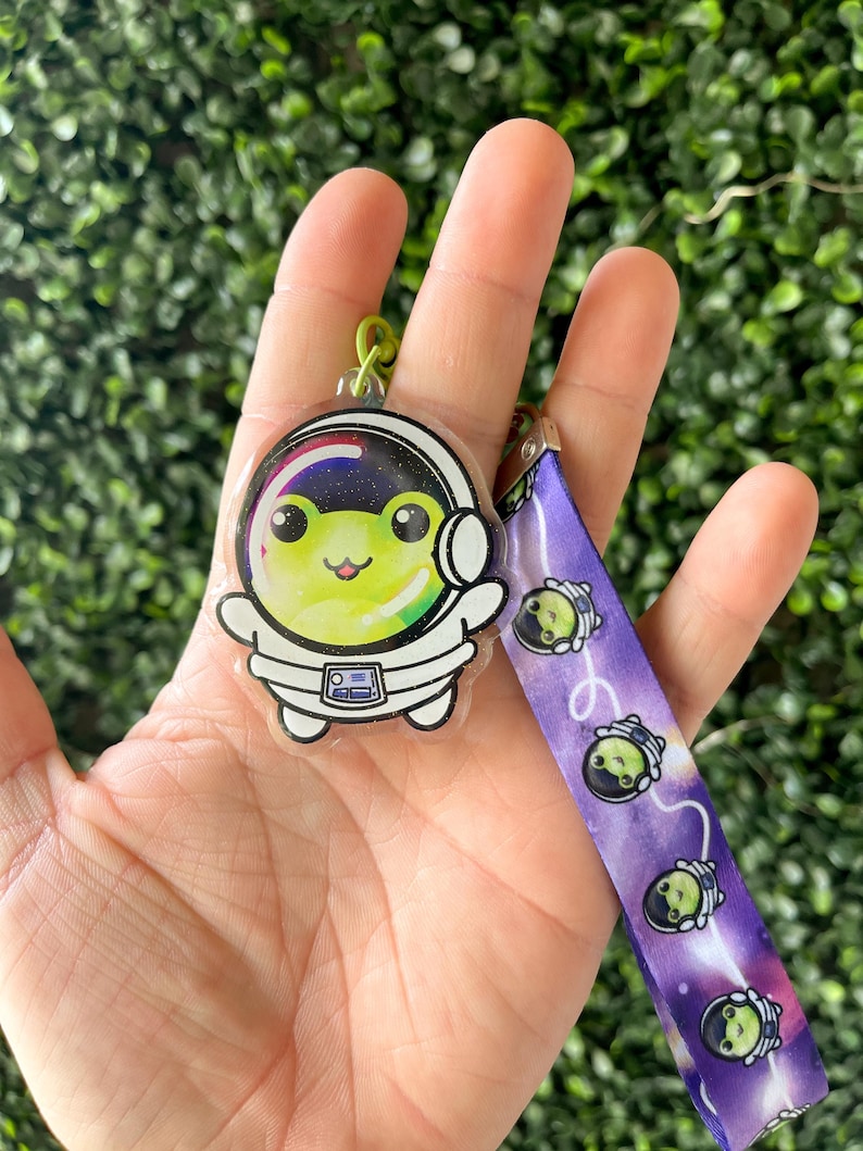 Pu&ograve; includere: Un portachiavi a forma di rana astronauta verde su sfondo galattico. Il portachiavi &egrave; attaccato a un laccio viola con un motivo ripetuto della rana astronauta.