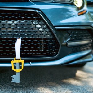 May include: A blue car with a key-shaped charm hanging from the front grille. The charm is yellow and gray and has a white strap.