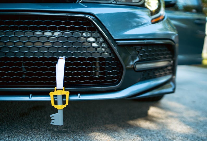 May include: A blue car with a key-shaped charm hanging from the front grille. The charm is yellow and gray and has a white strap.