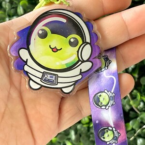 Pu&ograve; includere: Un portachiavi in plastica trasparente con una rana verde vestita da astronauta e un design a galassia viola e bianco. Il portachiavi &egrave; dotato di un laccio a galassia viola e bianco abbinato con un'immagine ripetuta della rana astronauta.