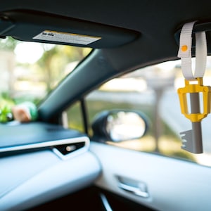 May include: A yellow and gray key-shaped car charm hanging from the rearview mirror of a car. The charm is attached to a white strap with a yellow button.