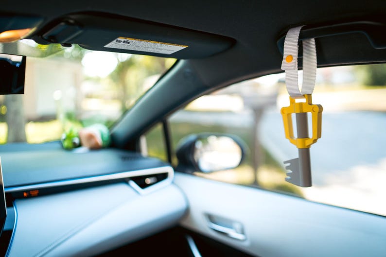 May include: A yellow and gray key-shaped car charm hanging from the rearview mirror of a car. The charm is attached to a white strap with a yellow button.