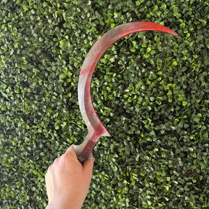 May include: A red and black sickle-shaped prop with a wooden handle. The prop is held in a hand against a green background.