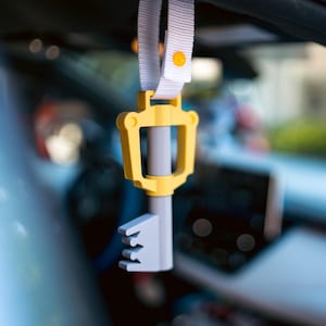 Op de afbeelding: Een gele en grijze plastic sleutelhanger in de vorm van een sleutel, hangend aan een wit bandje met een gele knop.