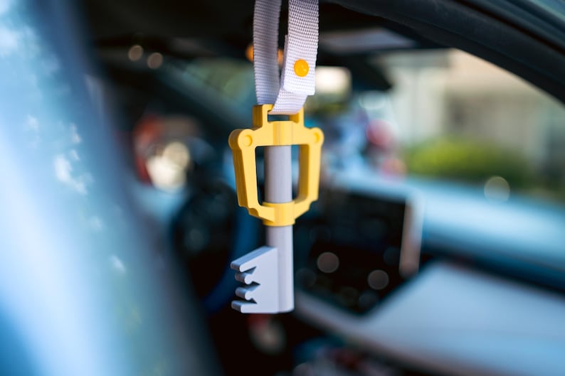 May include: A yellow and gray plastic key-shaped car charm hanging from a white strap with a yellow button.