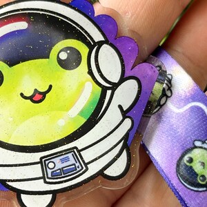 Pu&ograve; includere: Un ciondolo di rana astronauta verde brillante e trasparente con uno sfondo di galassia viola e bianco. La rana indossa una tuta da astronauta bianca con un casco nero e un pannello di controllo nero e bianco sul petto. Il ciondolo &egrave; attaccato a un nastro di galassia viola e bianco.