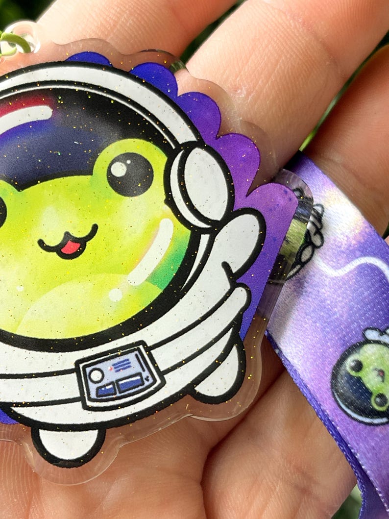 Pu&ograve; includere: Un ciondolo di rana astronauta verde brillante e trasparente con uno sfondo di galassia viola e bianco. La rana indossa una tuta da astronauta bianca con un casco nero e un pannello di controllo nero e bianco sul petto. Il ciondolo &egrave; attaccato a un nastro di galassia viola e bianco.