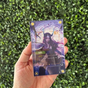 May include: A rectangular clear acrylic display case containing a collectible trading card featuring the character Irelia. The card displays purple and blue artwork, text details, and a power level of 5. The case is secured with four gold-colored screws at the corners.