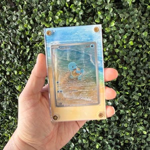 Pode incluir: Uma vitrine de acrílico transparente com um cartão colecionável Pokémon Squirtle. O cartão apresenta um personagem Squirtle azul em um fundo com tema de praia. A caixa é fixada com quatro parafusos dourados, e o fundo tem um gradiente azul e amarelo.