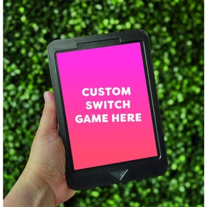 May include: A black electronic device with a pink and orange screen displaying the text "CUSTOM SWITCH GAME HERE." The device is held against a green, leafy background.