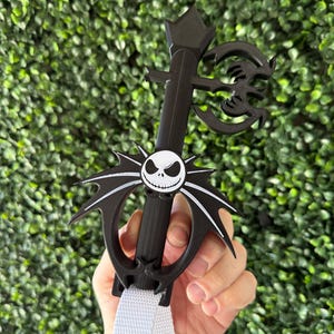 May include: Black plastic key chain shaped like a keyblade with a white Jack Skellington face on the front. The keyblade has a white strap with a black button.