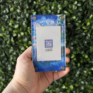 Puede incluir: Una vitrina acrílica azul y transparente con un inserto blanco y un código QR. La vitrina tiene tornillos dorados y un diseño abstracto colorido. El texto "PORGIS CORNER" es visible en el inserto.