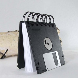 May include: A black floppy disk notepad with white paper pages, bound with black wire. The design includes a silver metal sliding cover and a silver metal centre piece. The notepad stands upright, a creative stationery item.