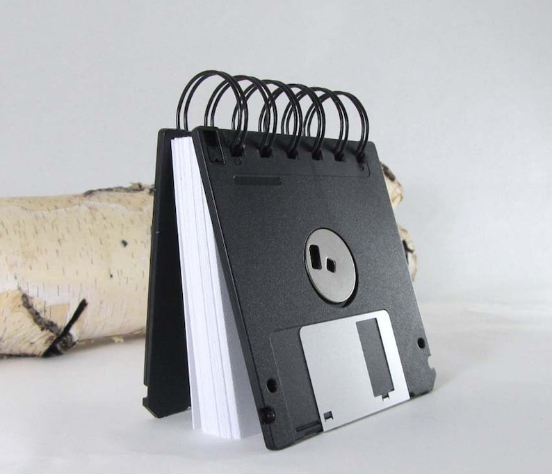 May include: A black floppy disk notepad with white paper pages, bound with black wire. The design includes a silver metal sliding cover and a silver metal centre piece. The notepad stands upright, a creative stationery item.